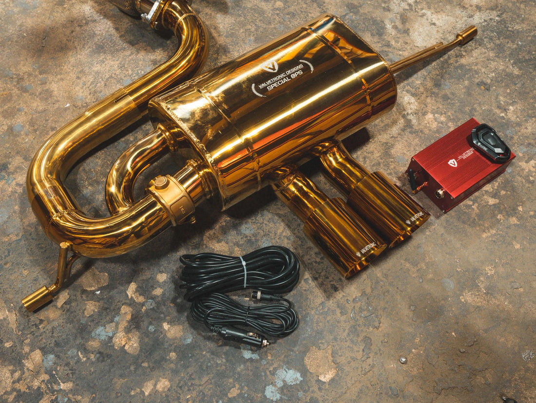 A close up view of Valvetronic Designs Anodized Gold Valved Sport Exhaust System's muffler, exhaust tips, valve controller and wires on the ground (for Lotus Evora).