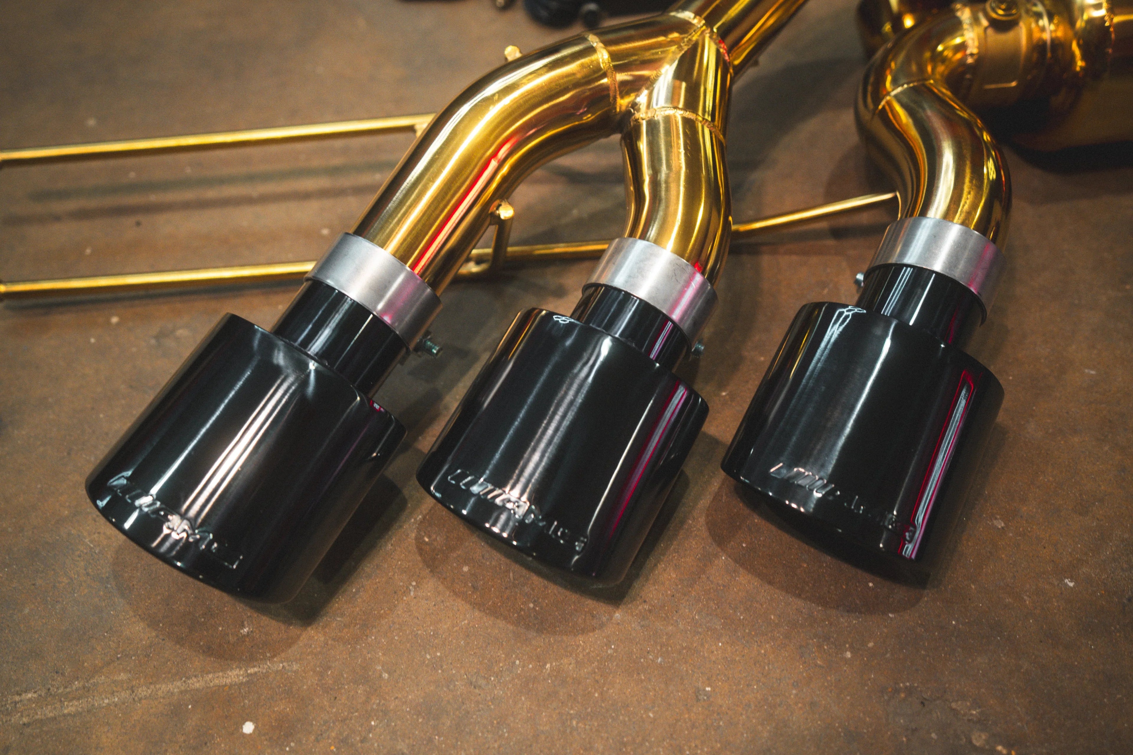 Close-up of the triple black exhaust tips connected to the polished gold-finish piping with silver clamps.