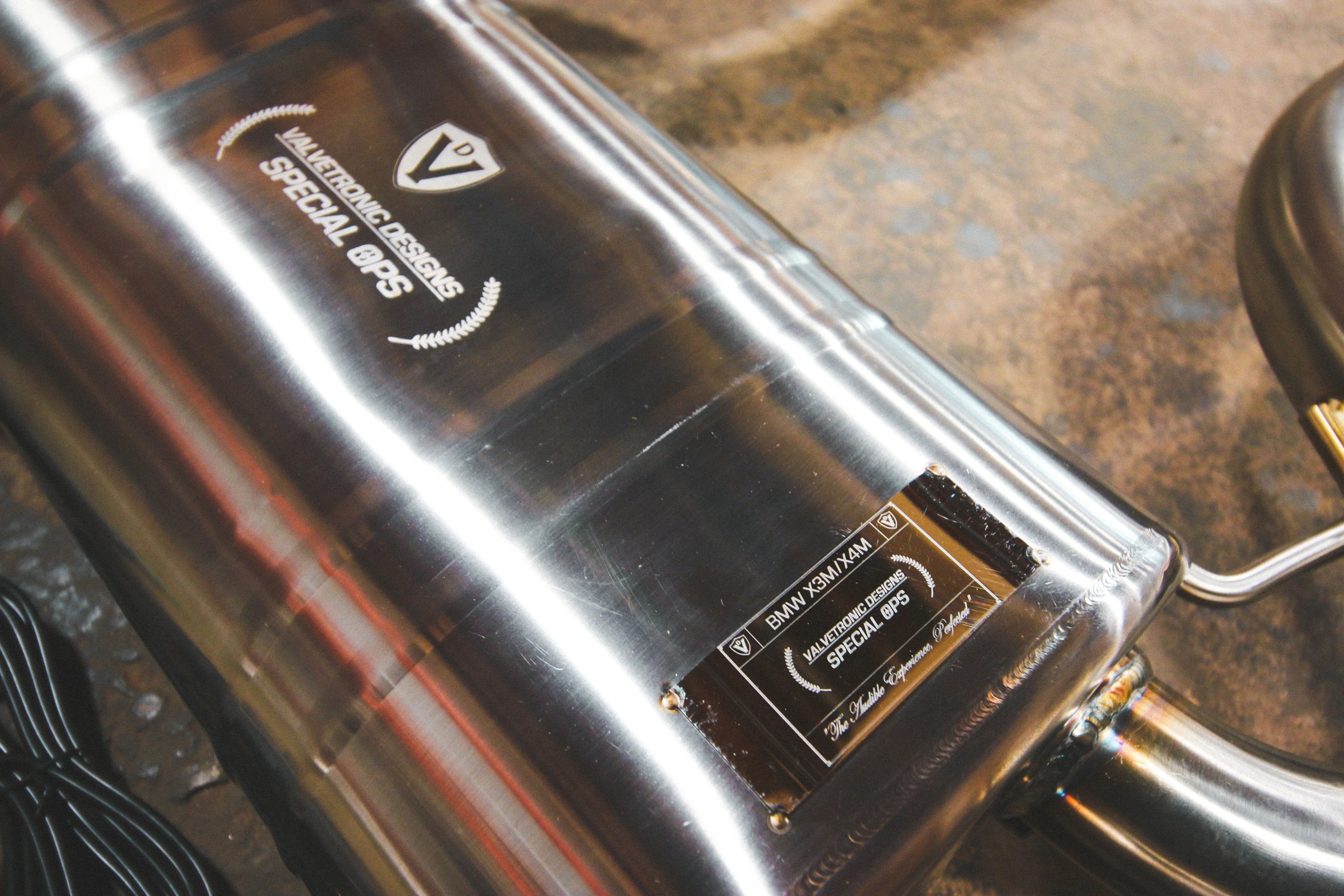A close-up of the rear muffler casing, featuring an engraved "Valvetronic Designs Special Ops" logo and a riveted identification plate marked for the BMW X3M/X4M.
