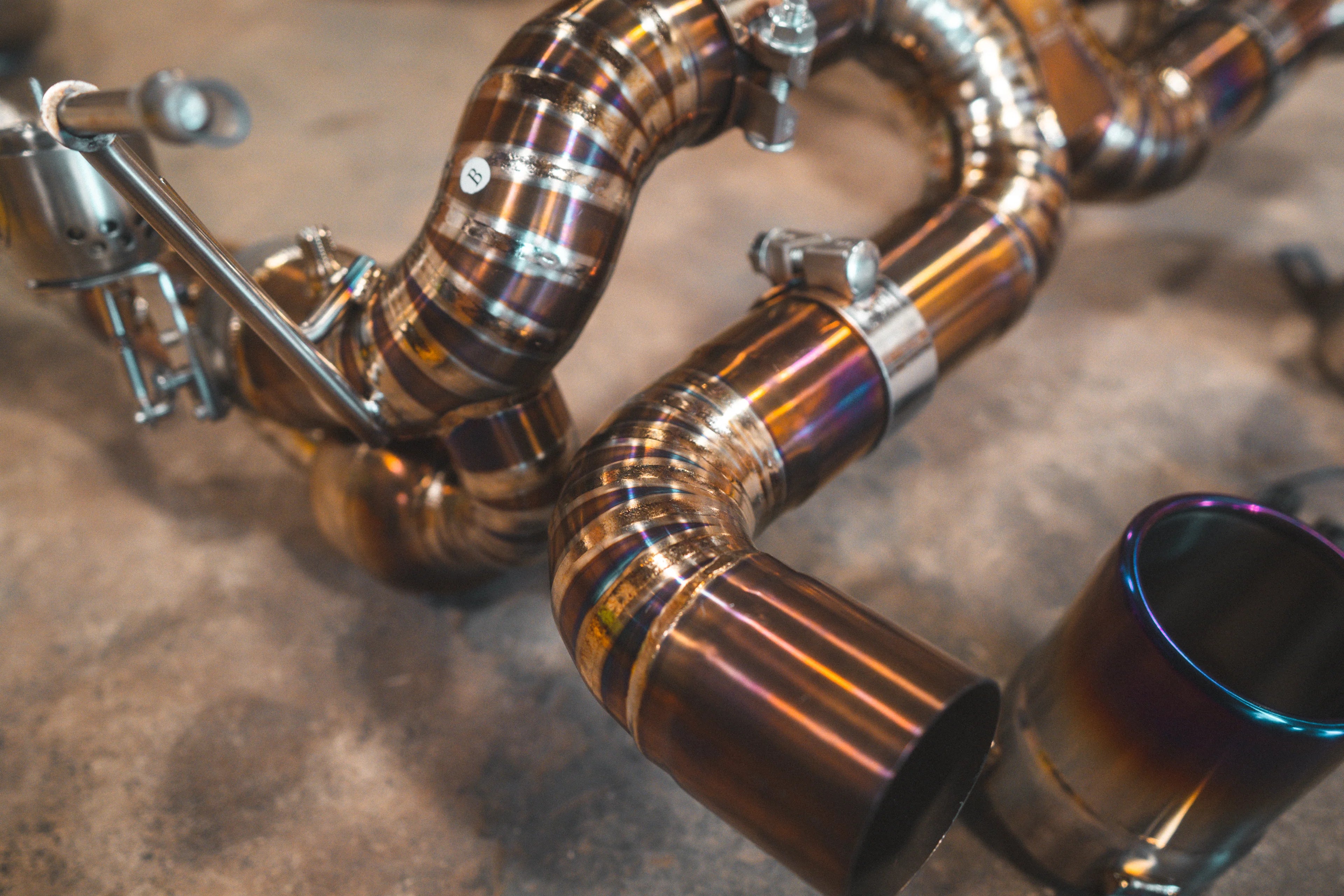 A close-up detail shot of the gold-finish exhaust pipe showing colorful heat discoloration at the weld site and a robotic weld bead.