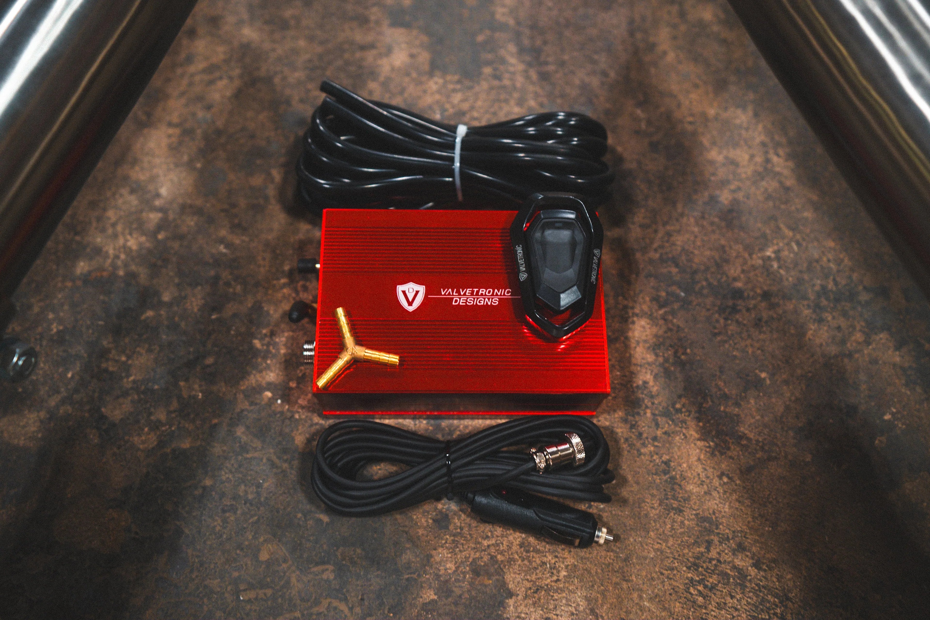 A red Valvetronic Designs valve control box shown with a remote, wiring harness, and vacuum fittings on a concrete surface.