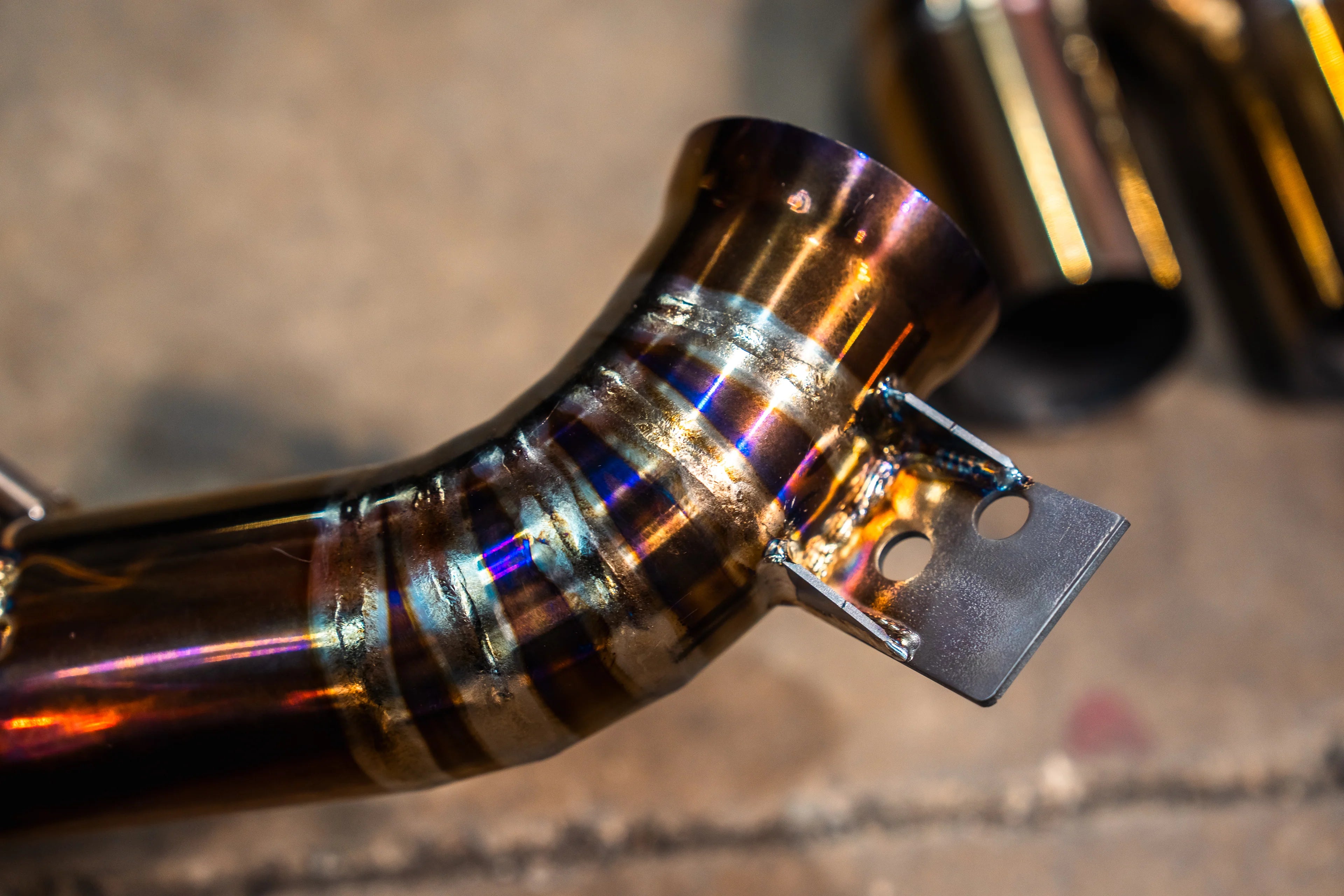 A close-up detail shot of a gold-finish exhaust pipe showing colorful heat discoloration and a robotic weld bead.