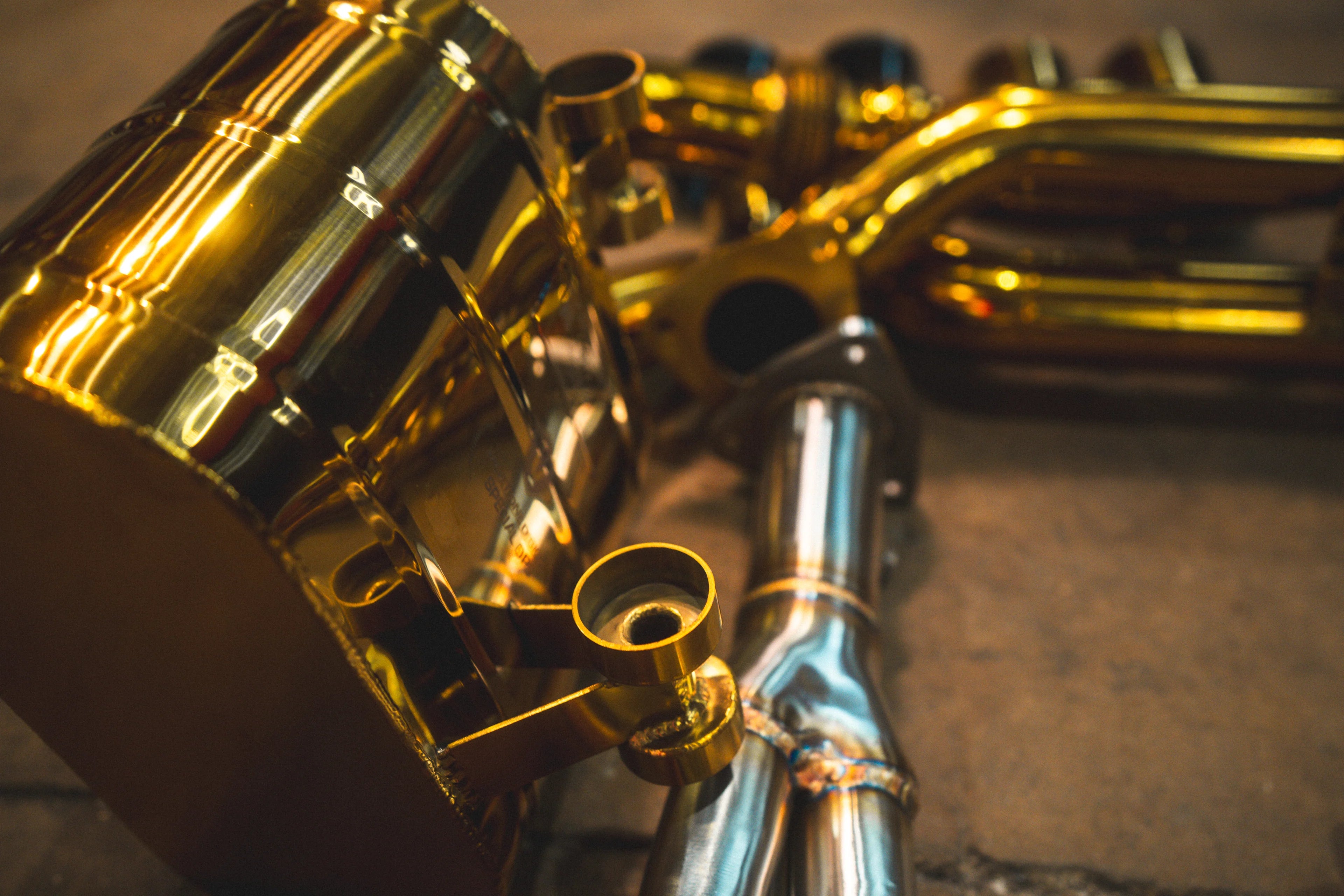 Macro shot of the gold-finish muffler casing, focusing on the mounting brackets and the precision TIG welding at the pipe junctions.