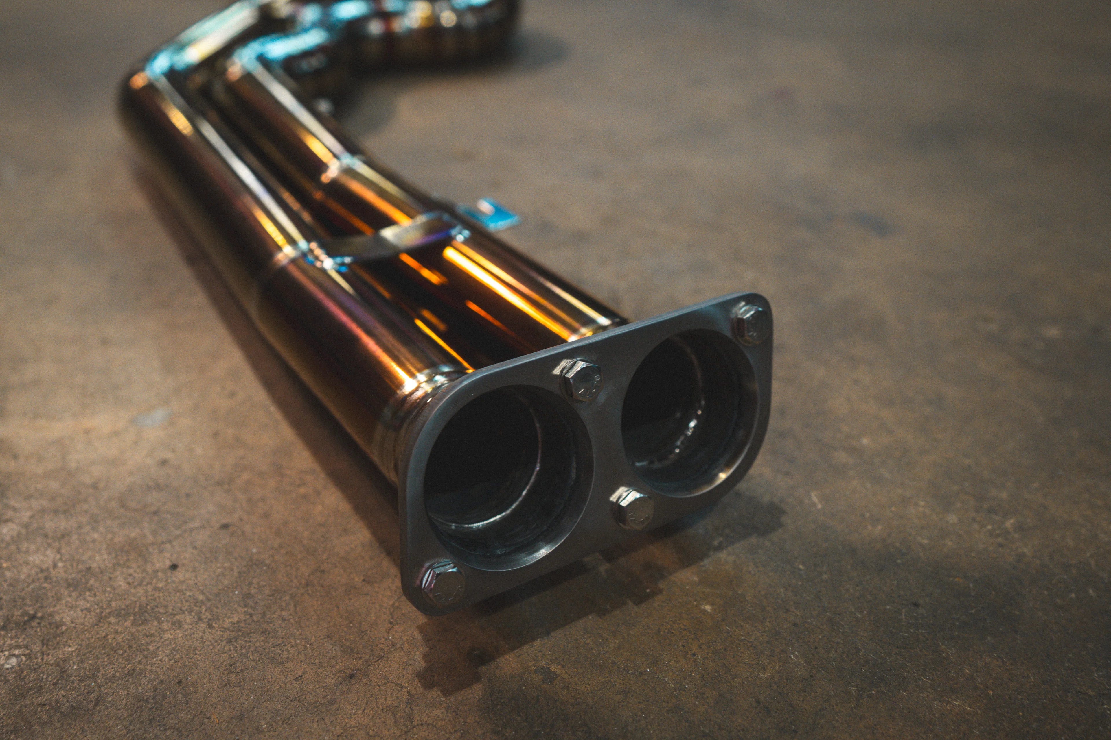 A close-up of the dual-inlet exhaust flange made of machined metal, showing the two circular openings and the bolted mounting points.