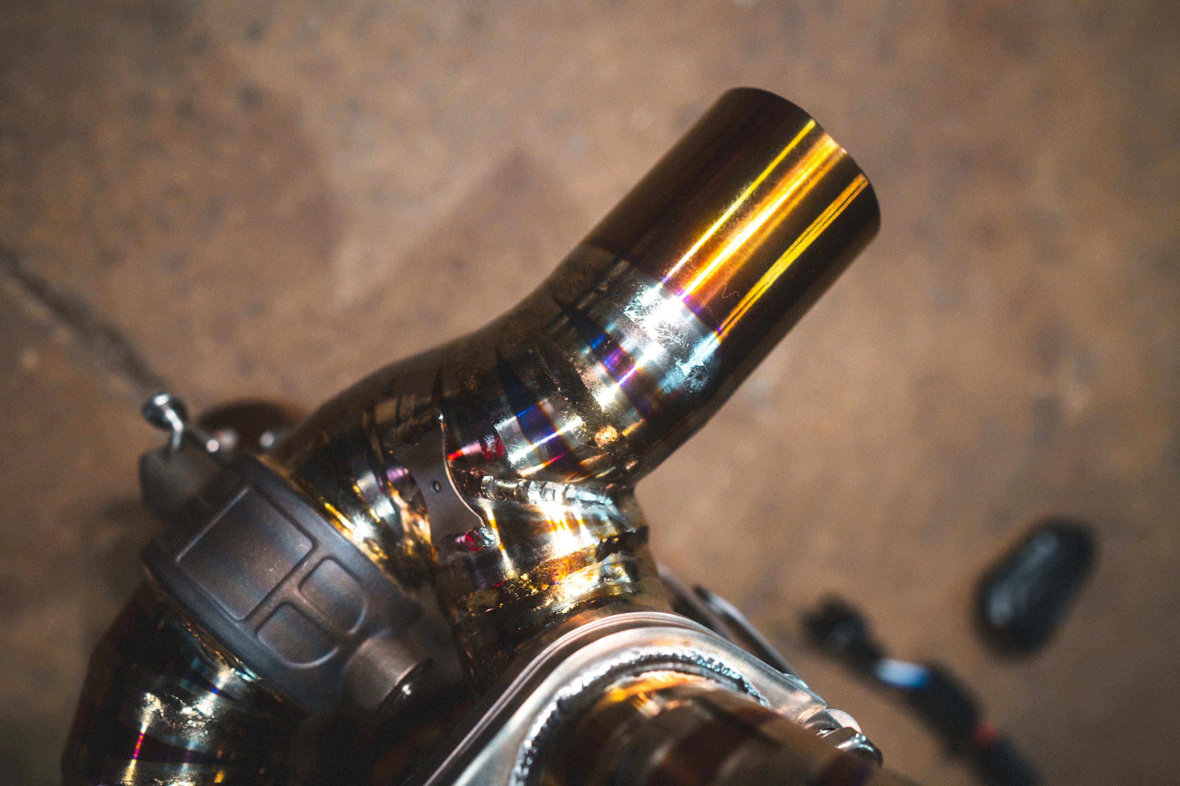 A close-up detail shot of a gold-finish exhaust pipe showing colorful heat discoloration and a robotic weld bead.
