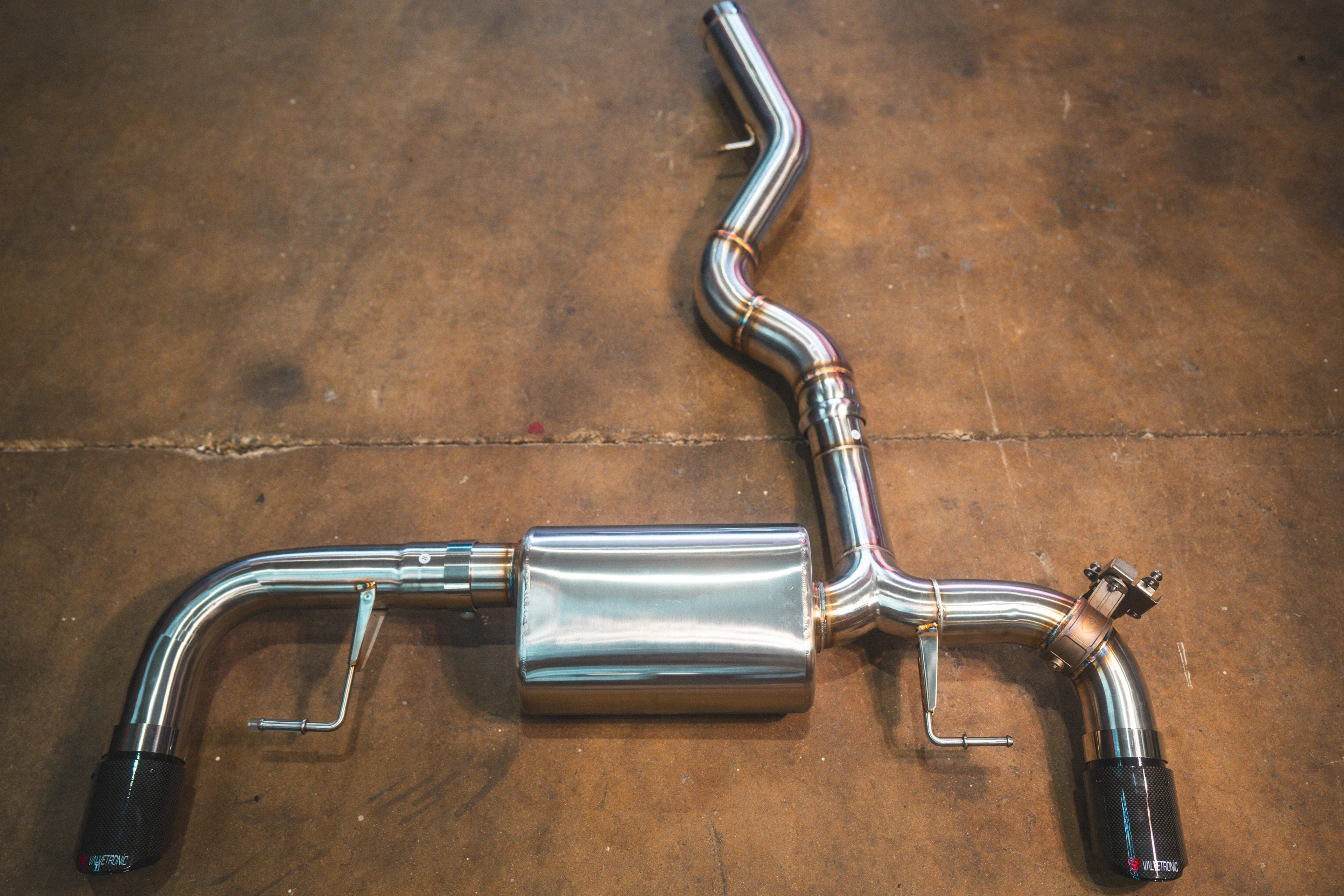 A stainless steel valved axle-back exhaust system with woven carbon fiber tips displayed on a concrete workshop floor.