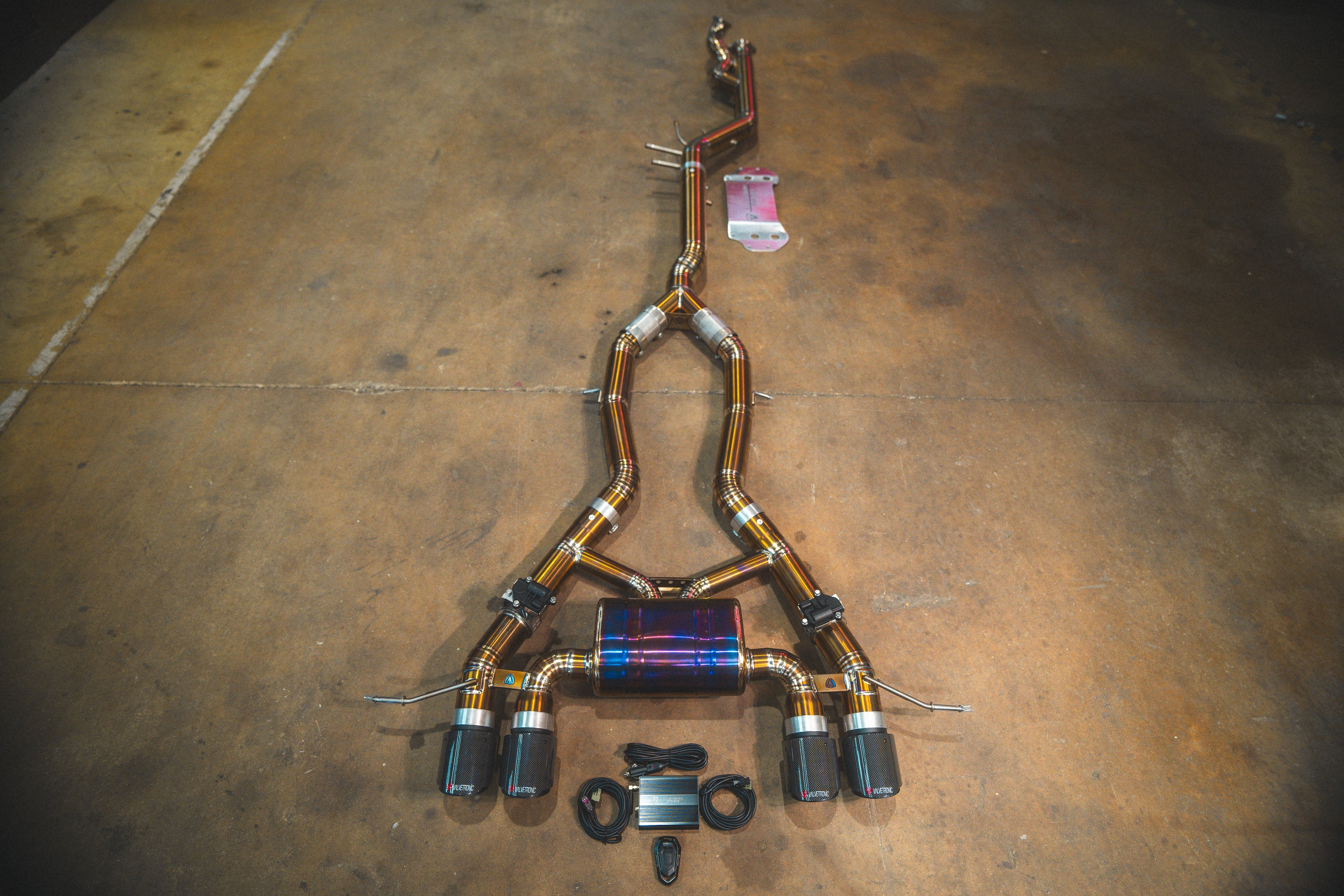 A full titanium valved exhaust system laid out on a concrete floor, featuring four traditional weave carbon fiber exhaust tips and a central purple/blue heat-treated muffler.