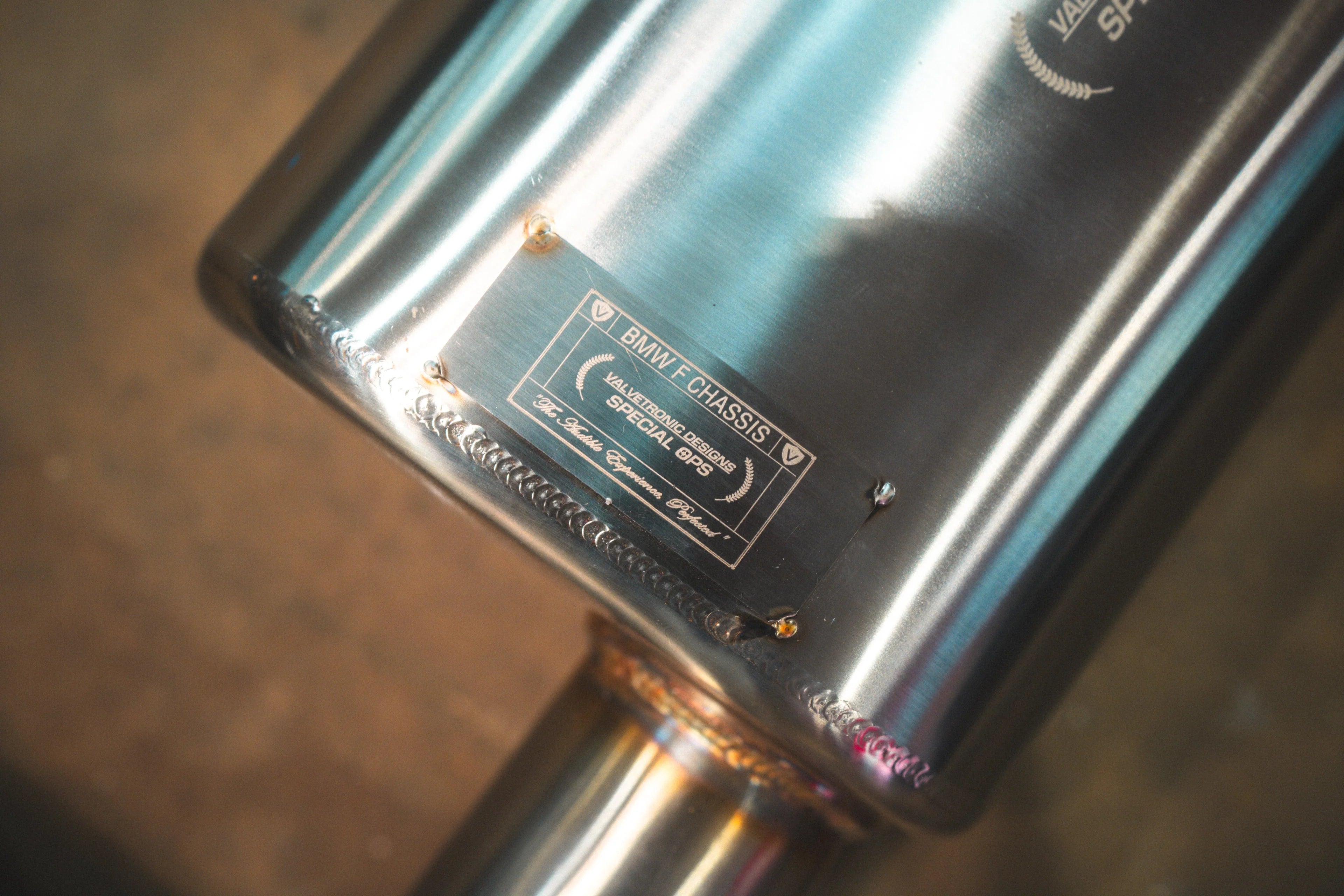 A close-up of the metal identification plaque welded to the muffler, which reads "BMW F CHASSIS - VALVETRONIC DESIGNS SPECIAL OPS".