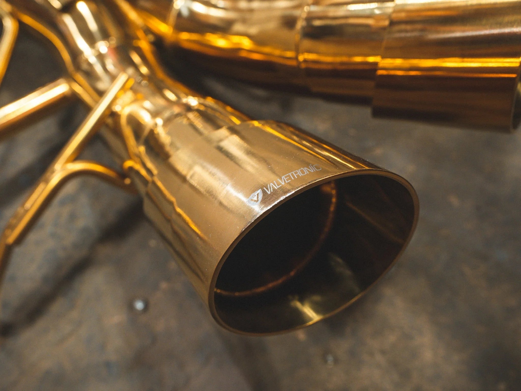Detailed close-up of a polished gold exhaust tip with the Valvetronic Designs logo etched on the rim.