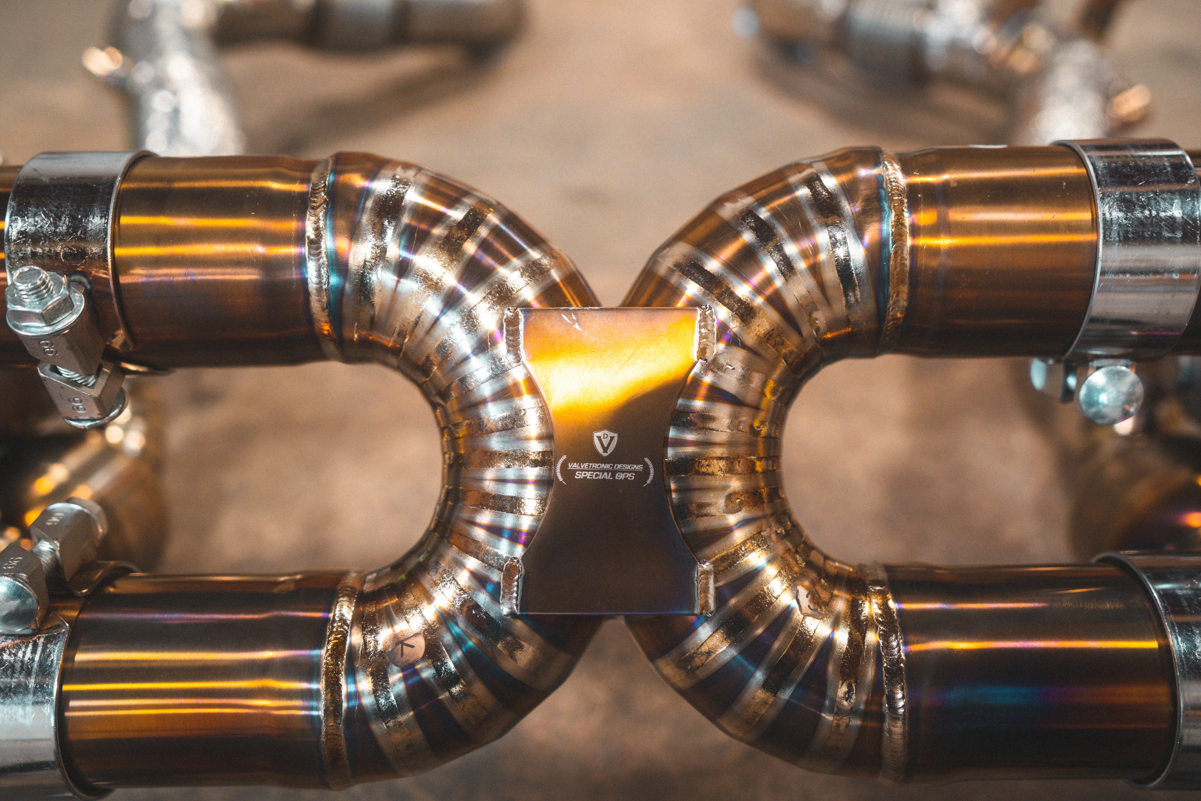 A macro shot focusing on the precision TIG welding and purple-blue heat tinting on a curved section of the gold exhaust piping.