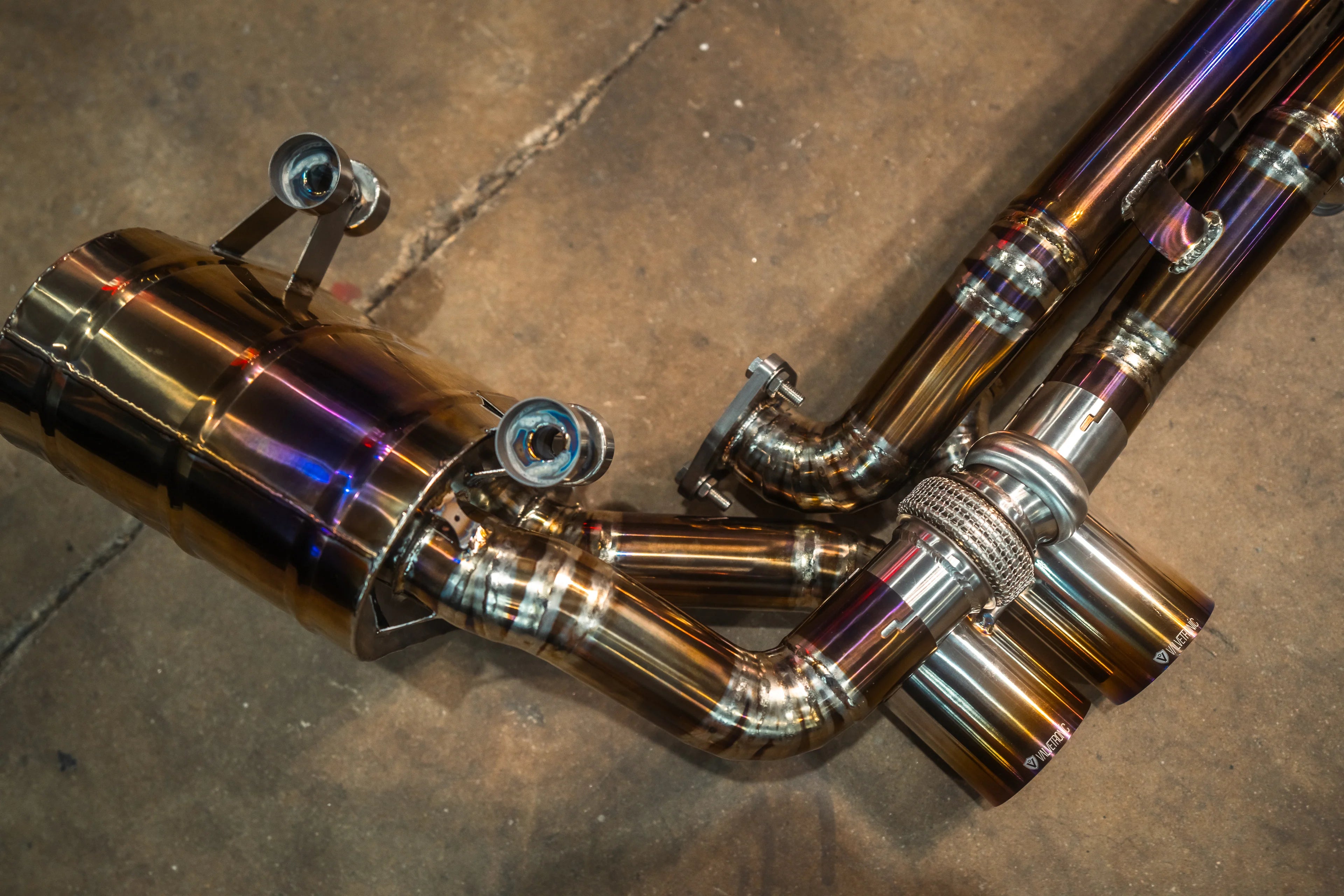 Detailed view of the titanium-finish muffler section, showing the electronic valve actuator, flexible mesh coupling, and the "Valvetronic" branded exhaust tips.