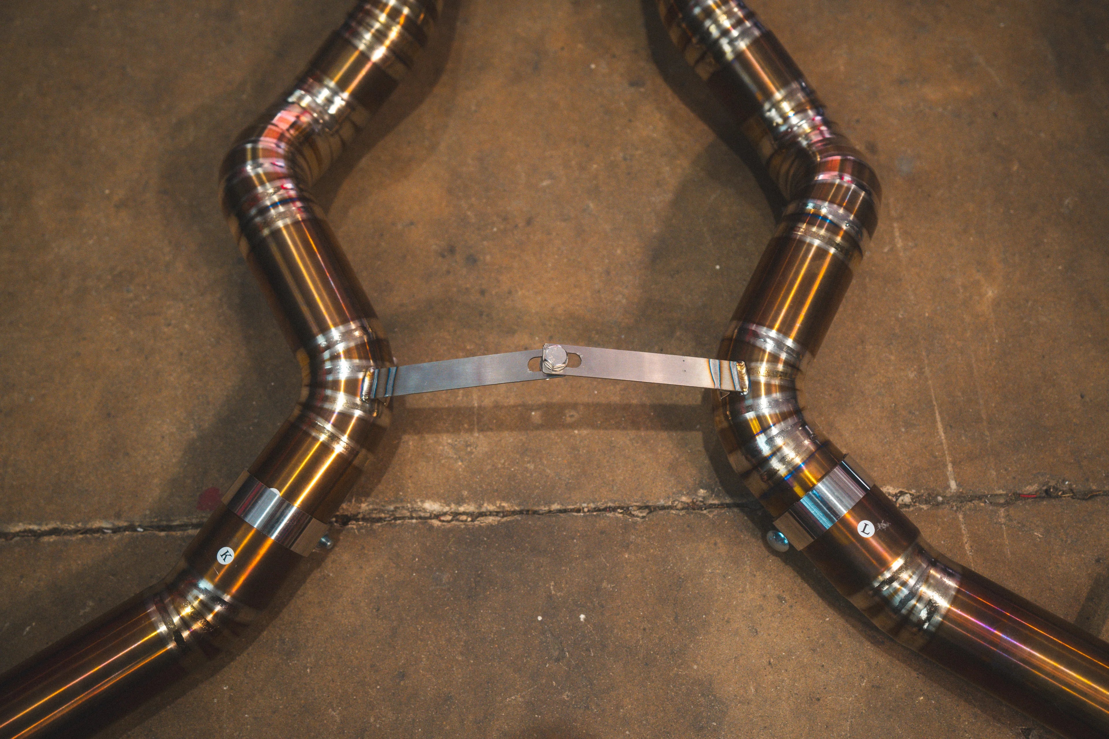 A close-up of the mid-pipe section showing a metal support bridge and circular band clamps connecting the titanium tubes.