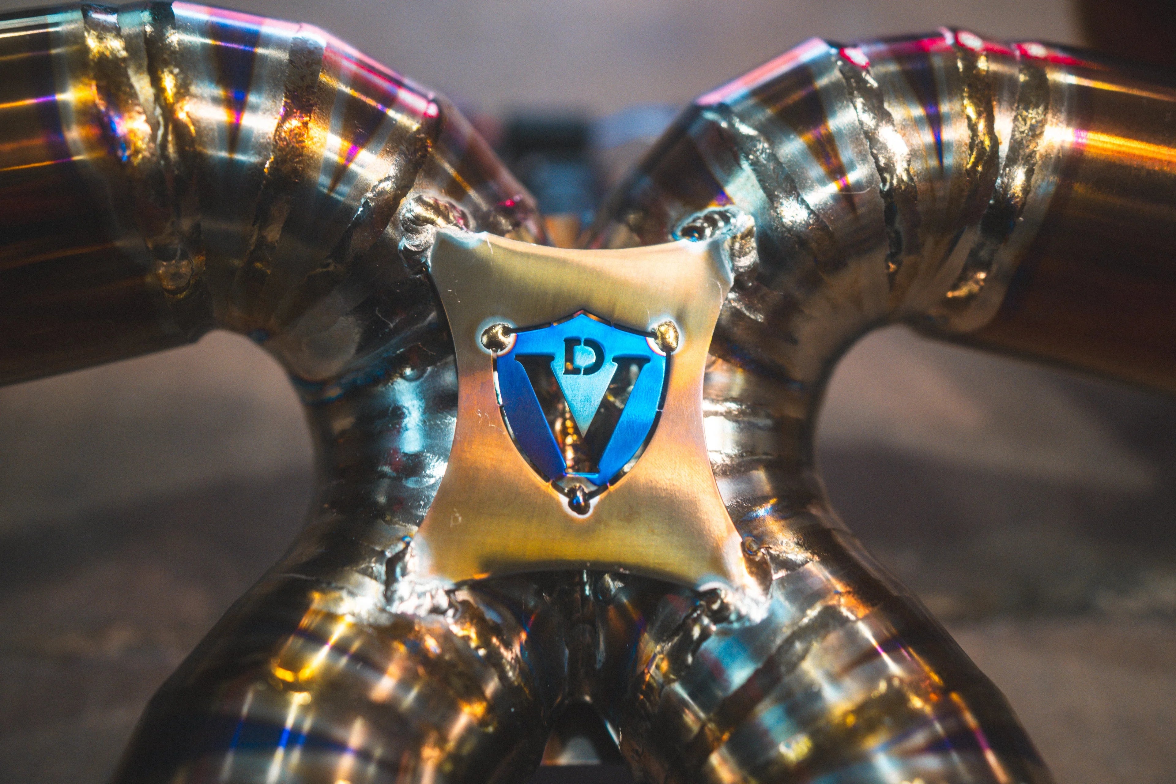 Macro shot of the Y-pipe junction, focusing on the blue-anodized Valvetronic "V" logo shield and the precision welding across the titanium-colored piping.