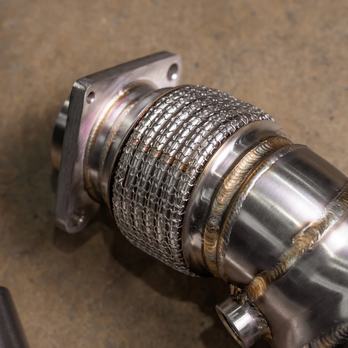 A close-up of a Valvetronic Designs 400 Cell Catted Downpipe focuses on the stainless steel interlock flex section and the thick mounting flange. 