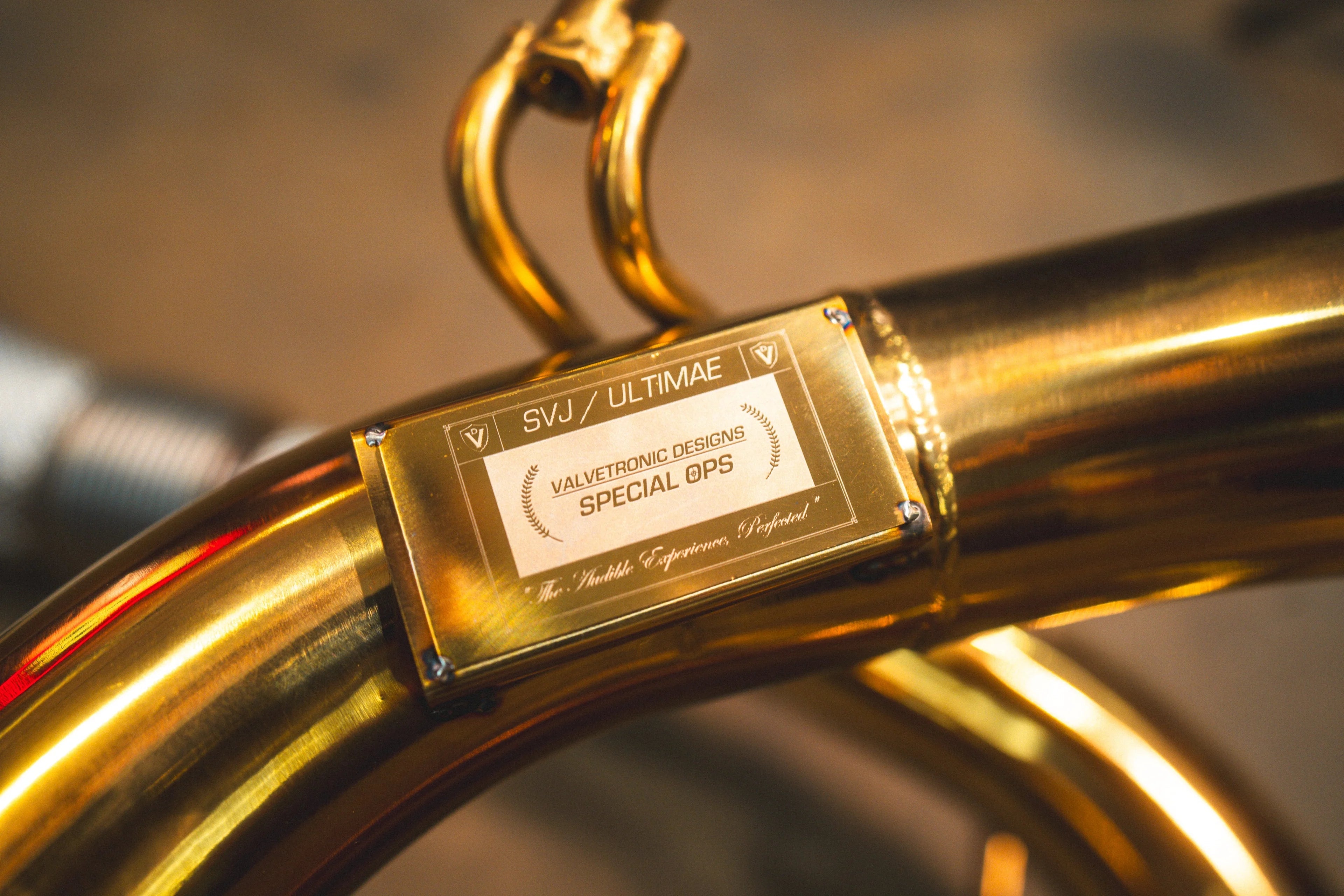 Extreme close-up of a gold-plated plaque welded to the exhaust pipe labeled "SVJ / ULTIMAE" and "Valvetronic Designs Special Ops."