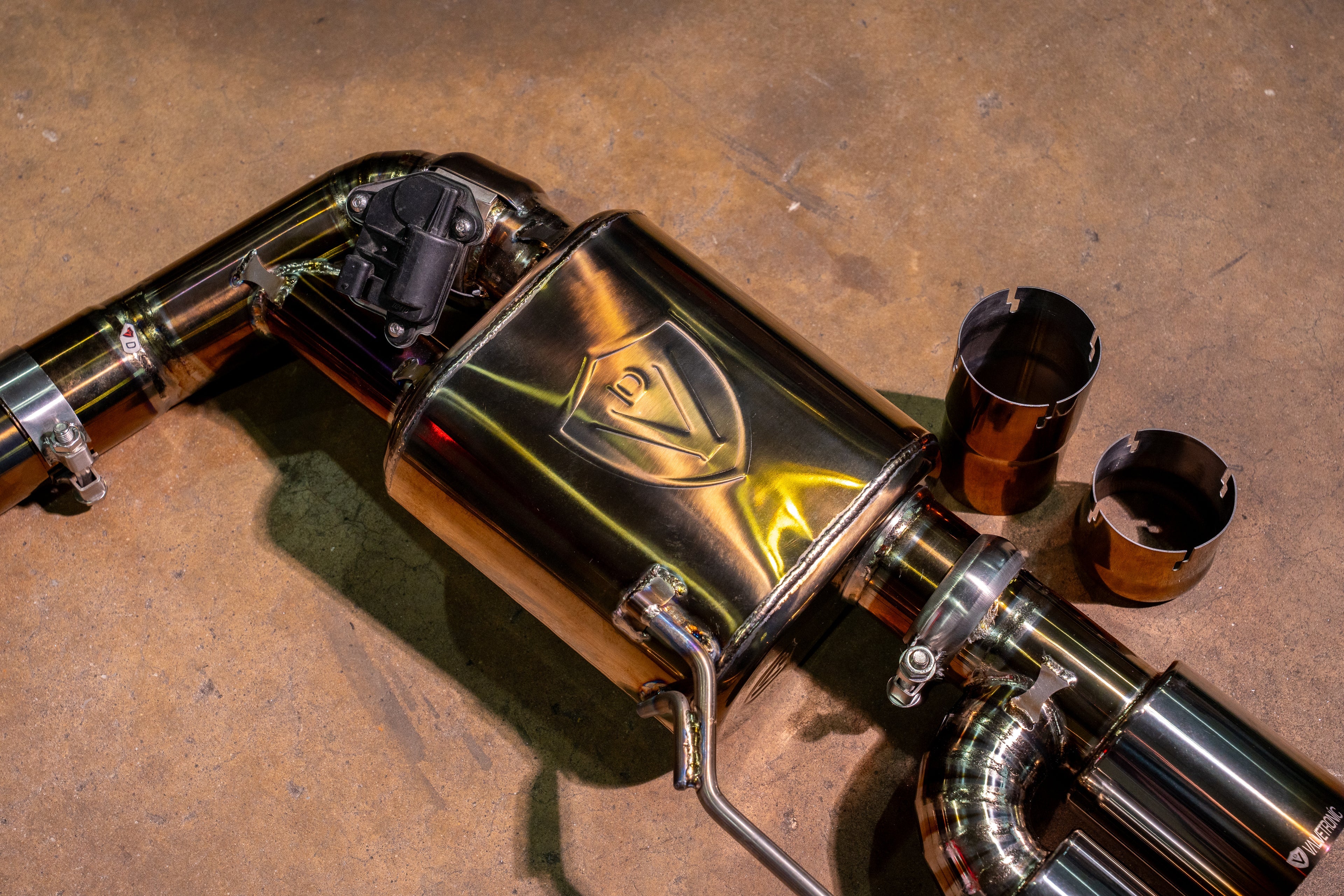 An angled close-up of the heat-treated muffler assembly, highlighting the embossed Valvetronic shield logo and the mounting bracket.