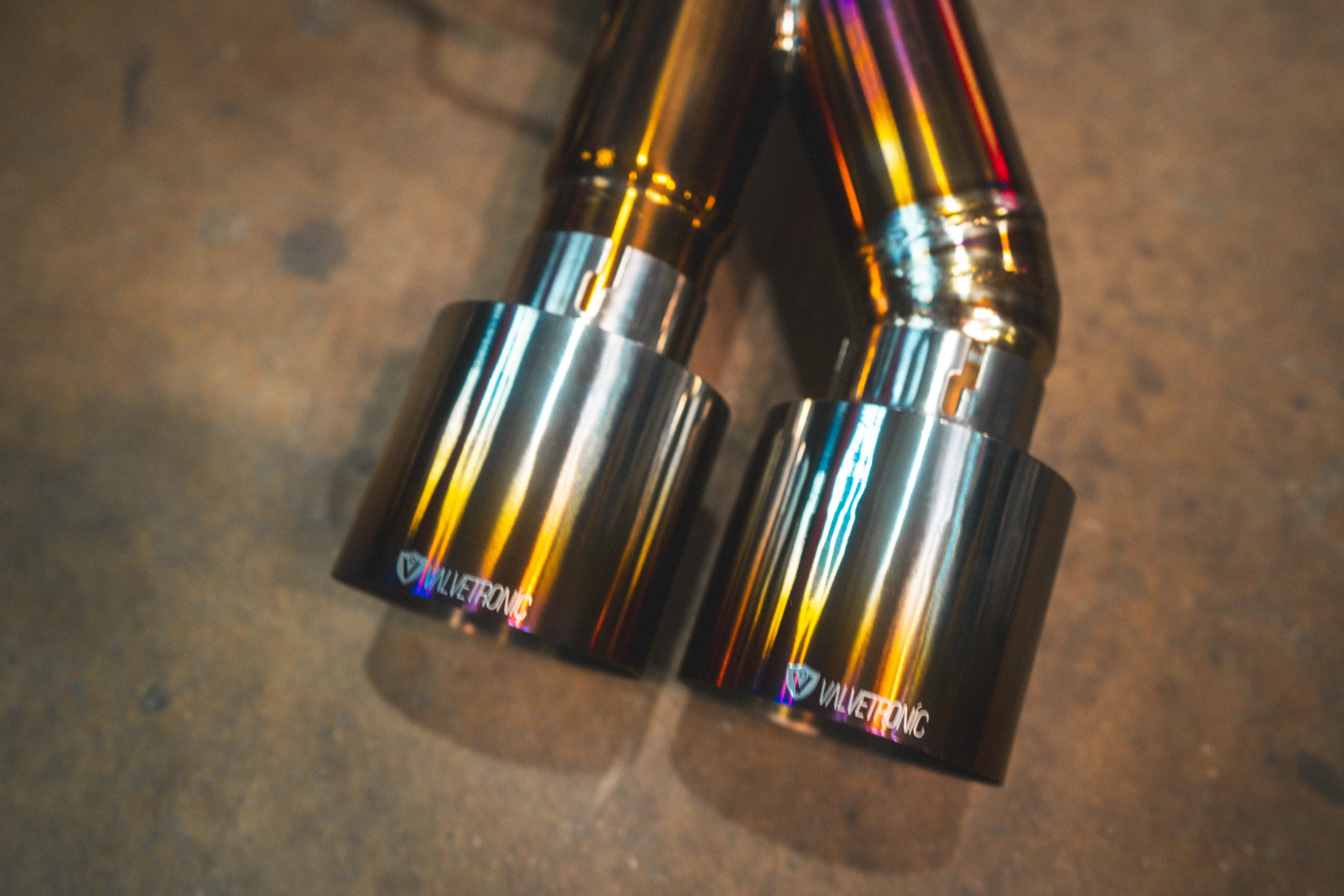 A close-up of two polished titanium exhaust tips, each featuring an engraved Valvetronic Designs logo and exhibiting a golden-purple iridescent finish.