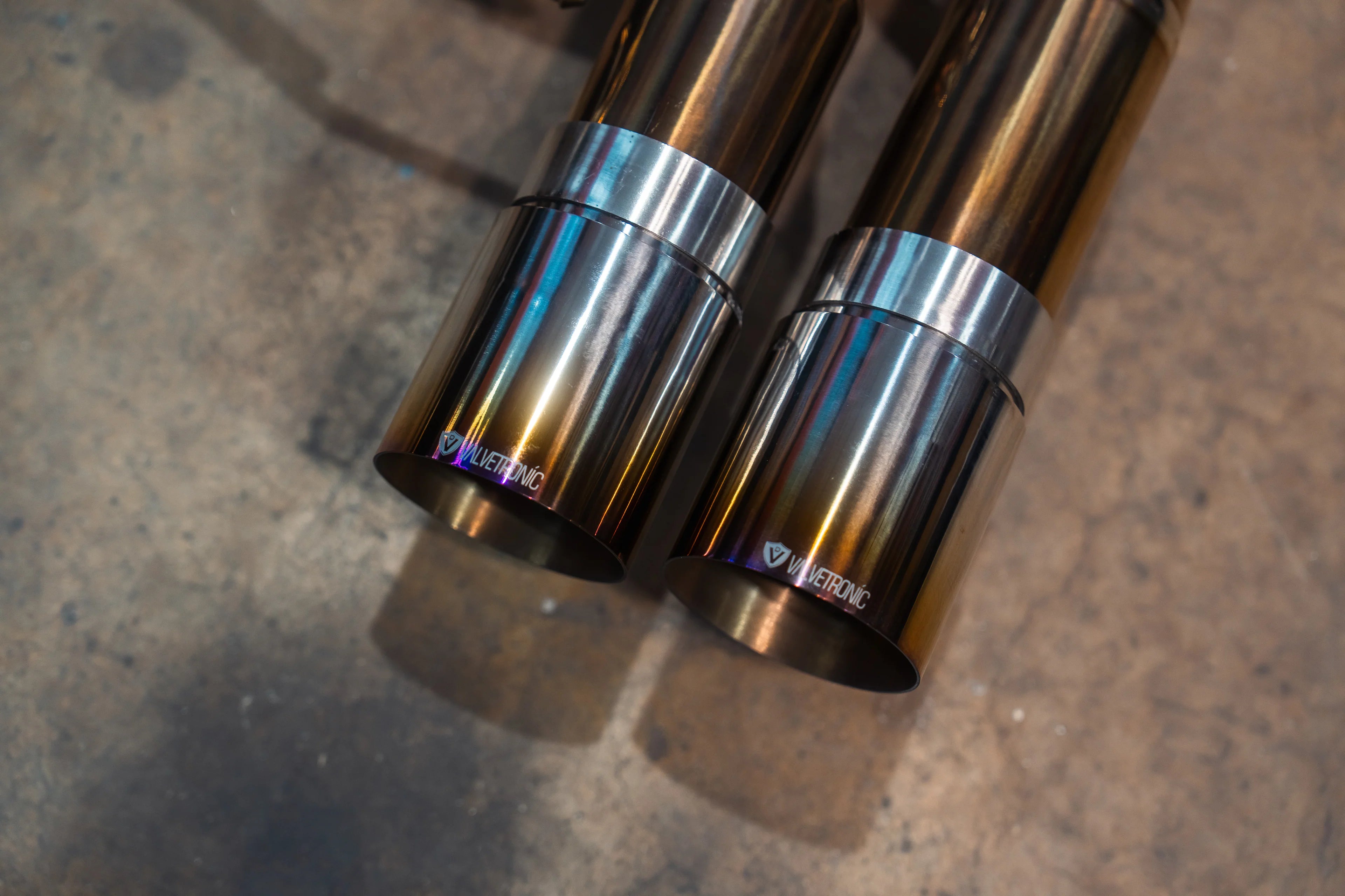 Detailed view of two burnt-finish titanium exhaust tips featuring the Valvetronic logo on a concrete workshop floor.