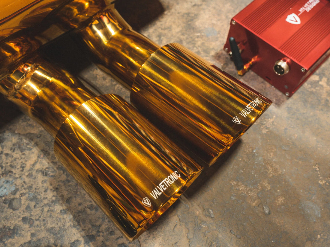 A close up view of Valvetronic Designs Anodized Gold Valved Sport Exhaust System's exhaust tips on the ground (for Lotus Evora).