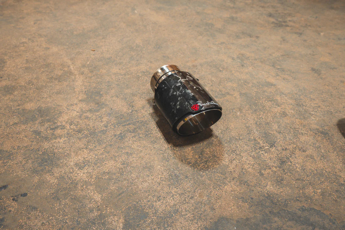 A side view of a single Valvetronic Designs Exhaust Tip in Forged Carbon Fiber on the ground.