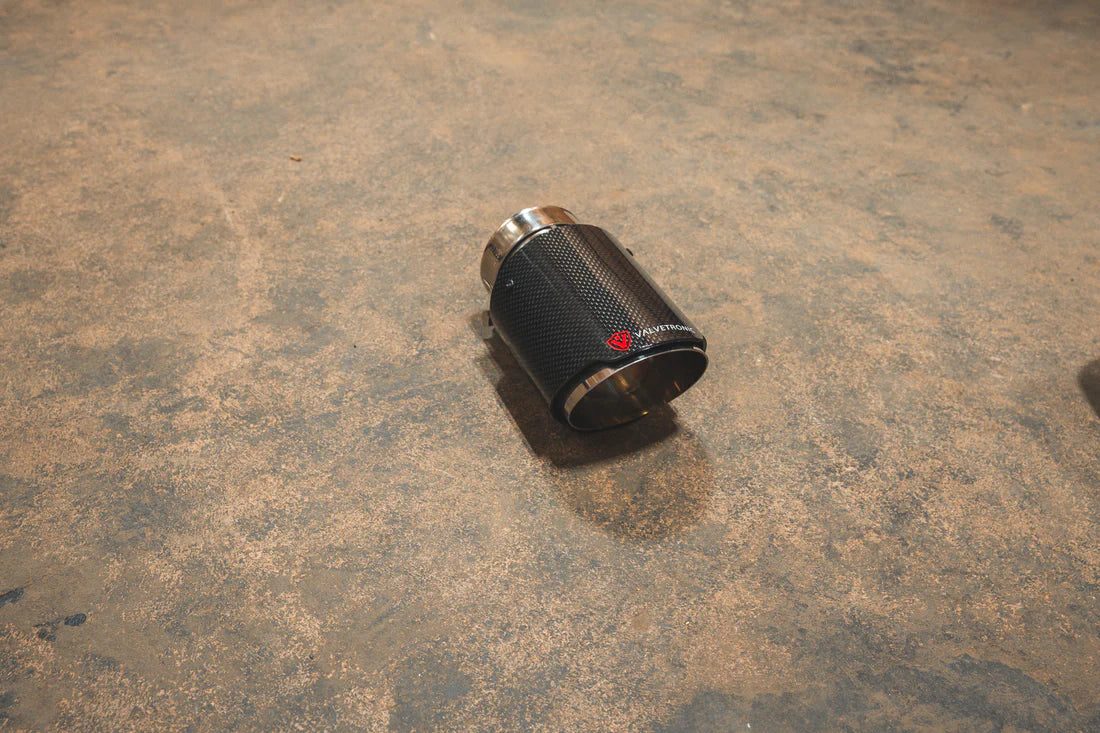 A side view of a single Valvetronic Designs Exhaust Tip in Carbon Fiber 1x1 on the ground.