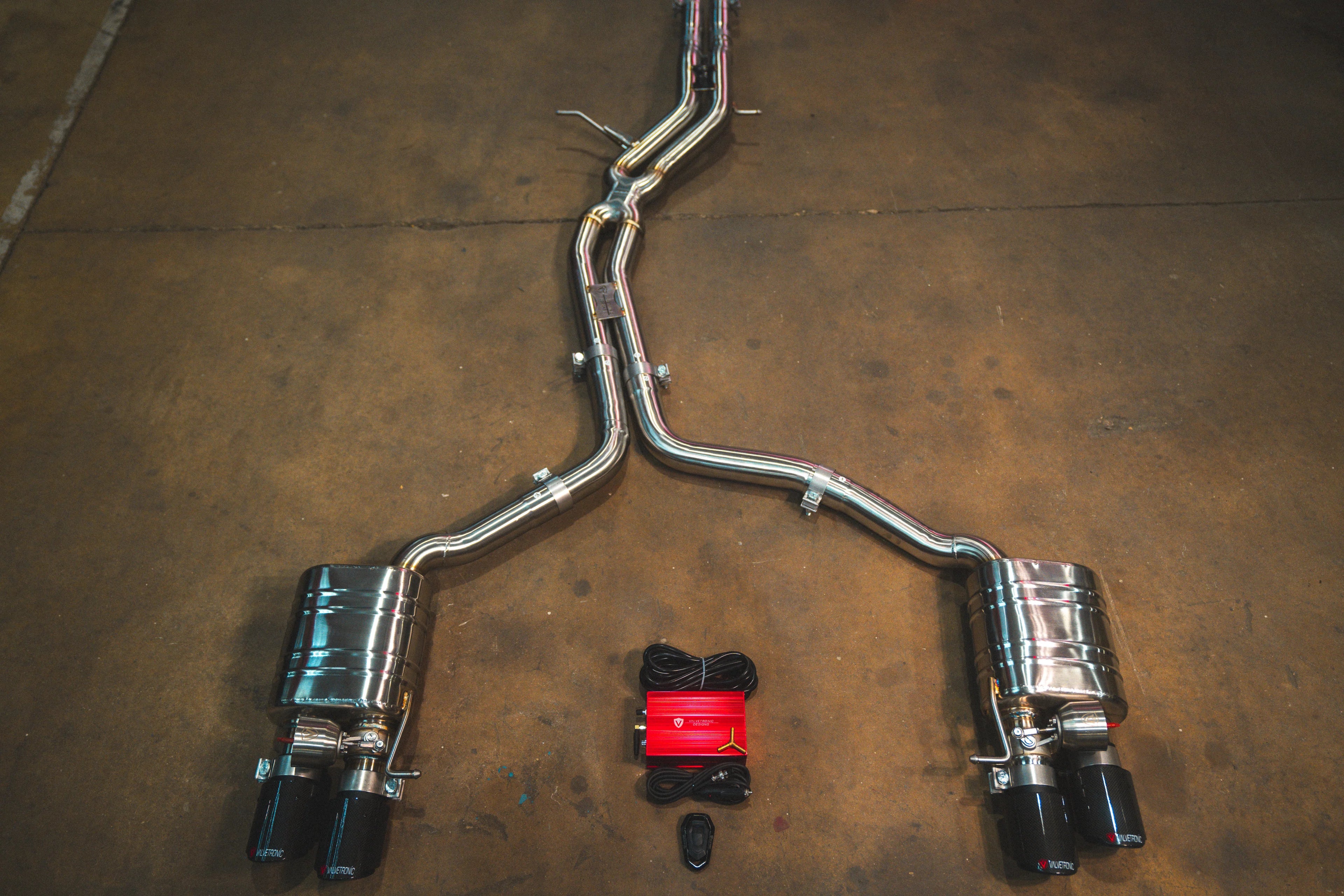 A full view of an stainless steel dual-exit exhaust system laid out on a concrete floor, featuring mid-pipes, two mufflers, carbon fiber exhaust tips, and accompanying wiring harnesses with a control box.