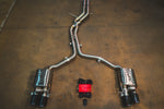A full view of an stainless steel dual-exit exhaust system laid out on a concrete floor, featuring mid-pipes, two mufflers, carbon fiber exhaust tips, and accompanying wiring harnesses with a control box.