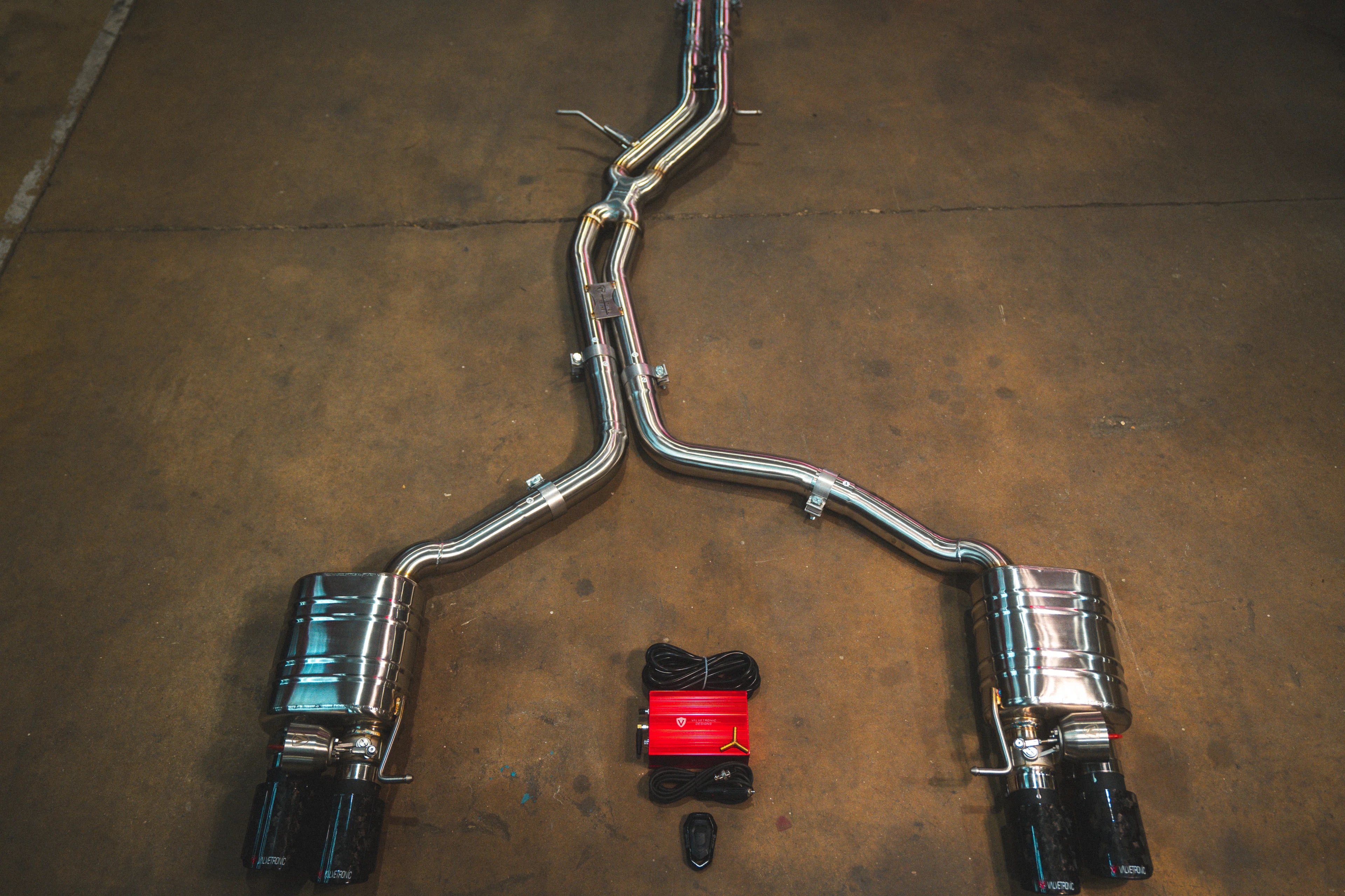 A full view of an stainless steel dual-exit exhaust system laid out on a concrete floor, featuring mid-pipes, two mufflers, forged carbon fiber exhaust tips, and accompanying wiring harnesses with a control box.