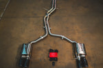 A full view of an stainless steel dual-exit exhaust system laid out on a concrete floor, featuring mid-pipes, two mufflers, forged carbon fiber exhaust tips, and accompanying wiring harnesses with a control box.