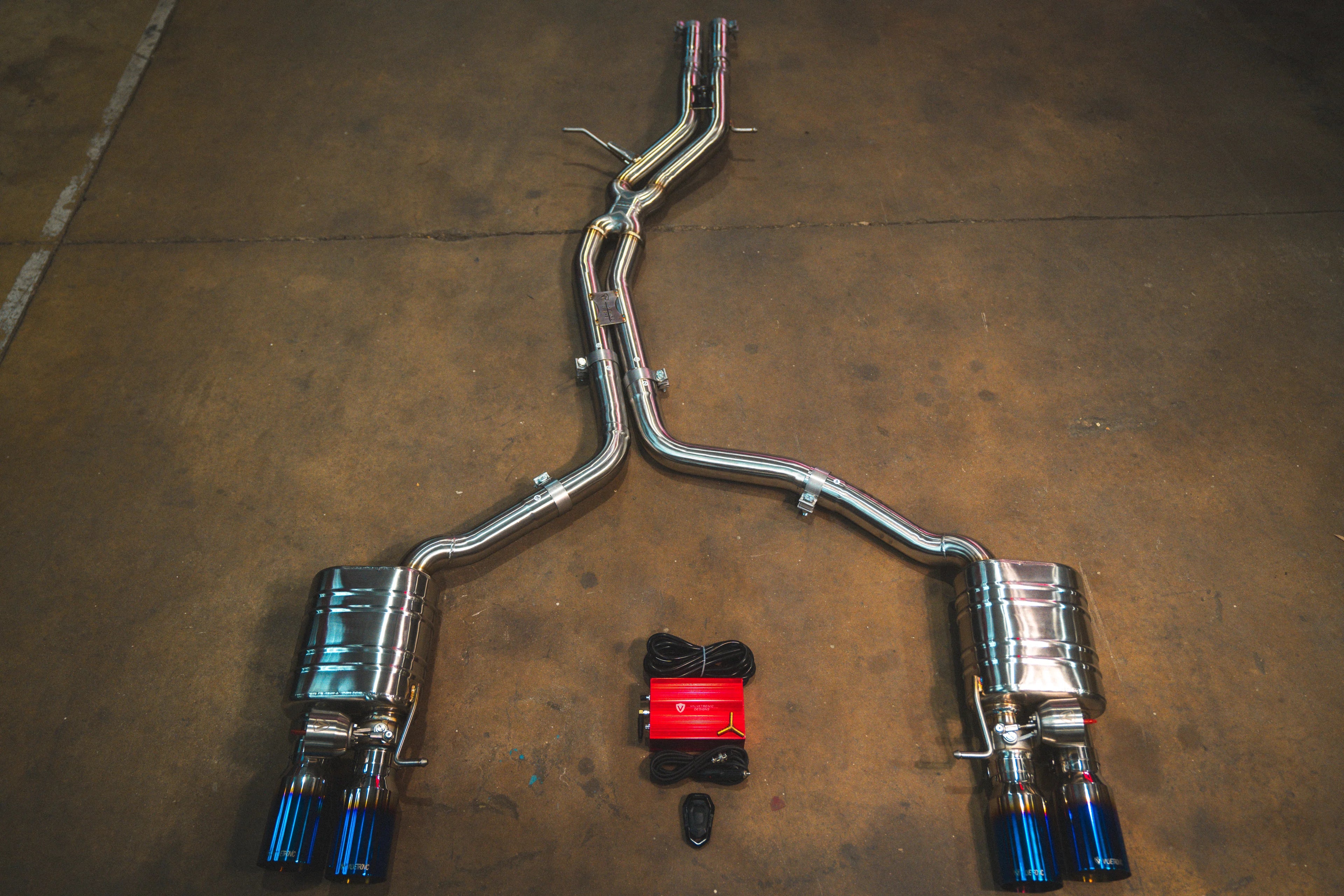 A full view of an stainless steel dual-exit exhaust system laid out on a concrete floor, featuring mid-pipes, two mufflers, burnt exhaust tips, and accompanying wiring harnesses with a control box.