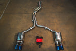 A full view of an stainless steel dual-exit exhaust system laid out on a concrete floor, featuring mid-pipes, two mufflers, burnt exhaust tips, and accompanying wiring harnesses with a control box.