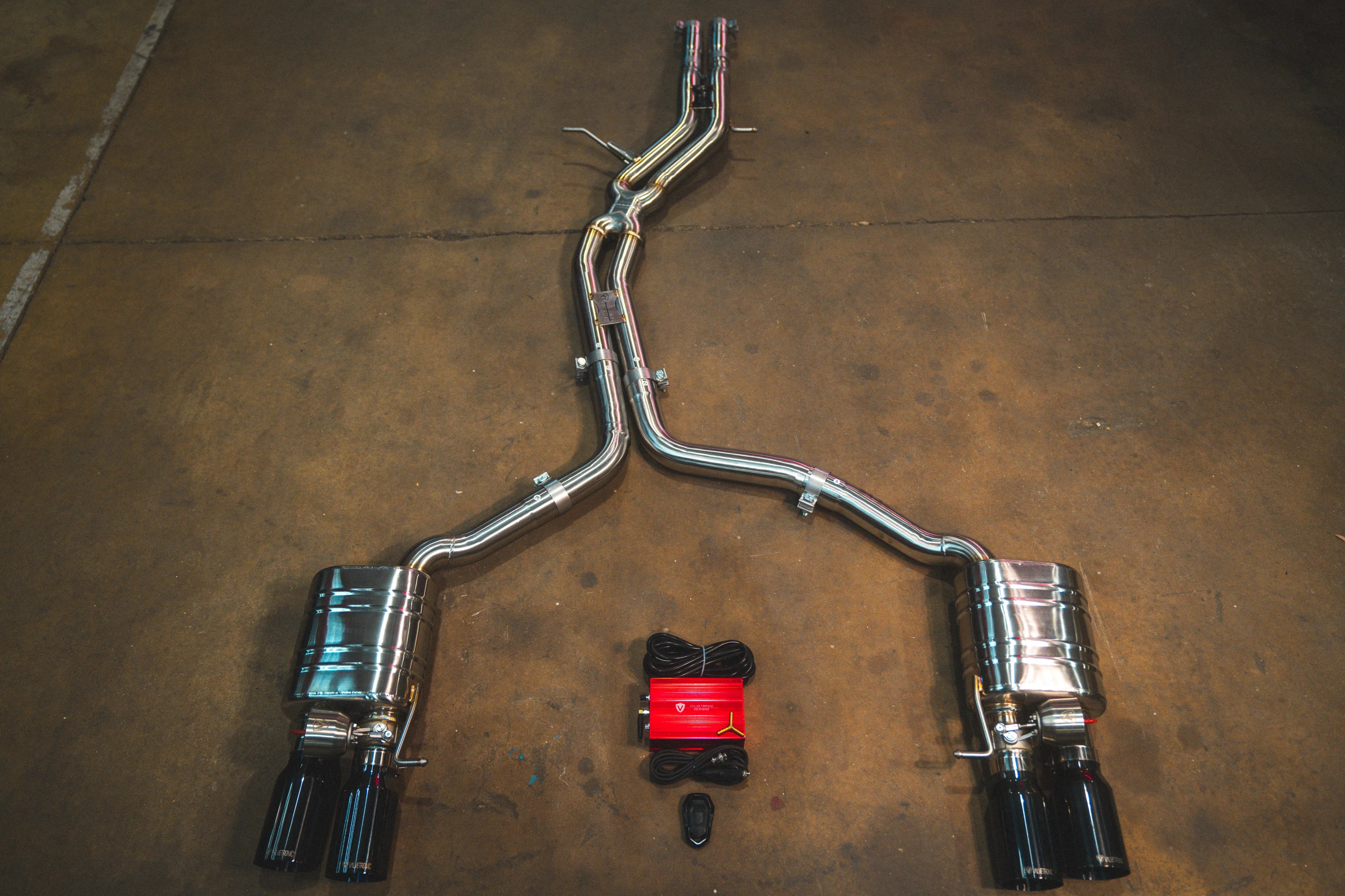 A full view of an stainless steel dual-exit exhaust system laid out on a concrete floor, featuring mid-pipes, two mufflers, black exhaust tips, and accompanying wiring harnesses with a control box.