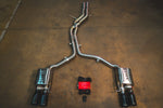 A full view of an stainless steel dual-exit exhaust system laid out on a concrete floor, featuring mid-pipes, two mufflers, black exhaust tips, and accompanying wiring harnesses with a control box.