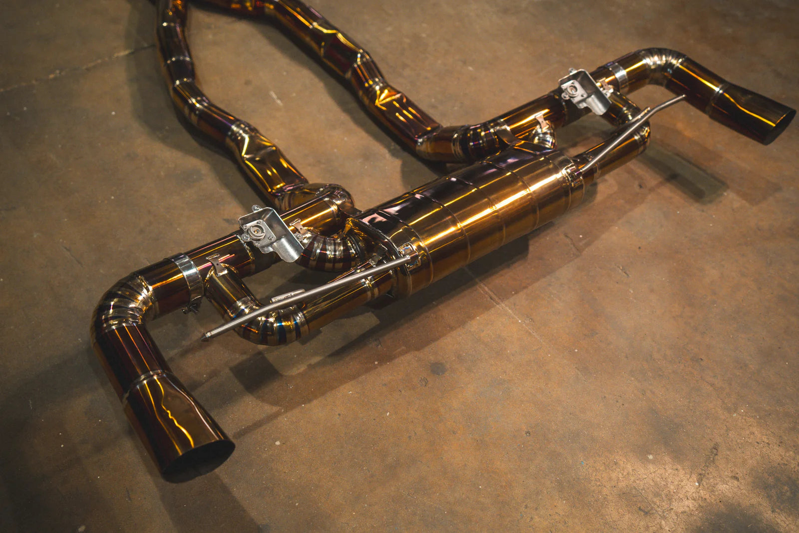 An angled, close-up view of the rear section of the Valvetronic Designs exhaust. The image highlights the polished gold-finish muffler and the two electronic valve controllers mounted on the piping, showcasing the intricate pie-cut welds and the iridescent purple hues of the titanium.