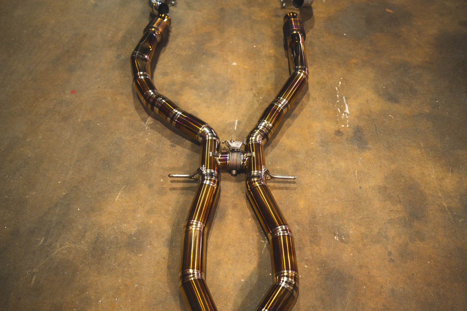 A detailed close-up of the exhaust system's X-pipe crossover. The image focuses on the high-quality TIG welding and the vibrant heat-treated coloring of the titanium. Small mounting brackets and a center support brace are visible against the concrete floor background.