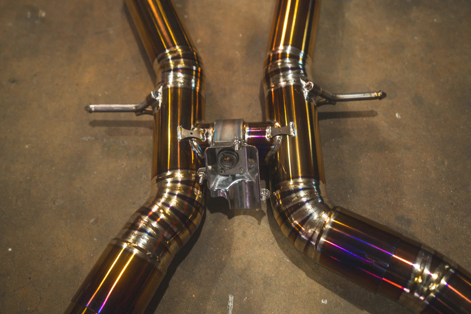 A top-down view of the midsection of the Valvetronic Designs exhaust system. This shot emphasizes the symmetrical flow of the twin titanium pipes as they merge into the center X-section, highlighting the consistent gold-anodized finish and professional weld patterns.