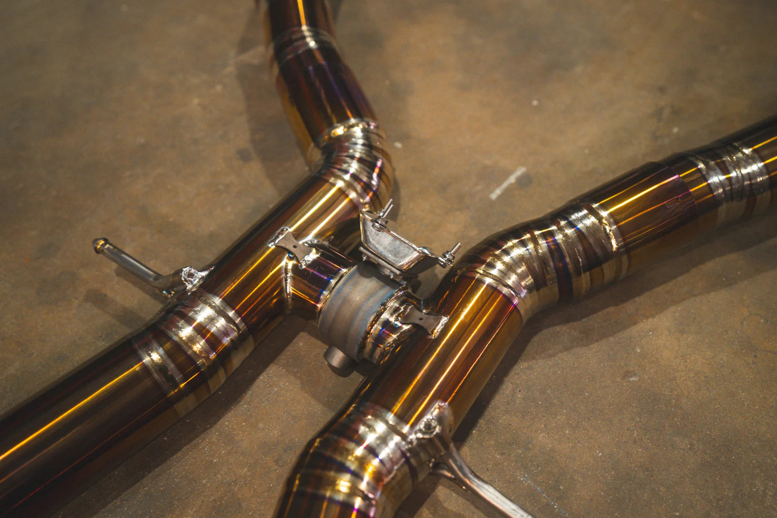 A macro shot focusing on an electronic valve actuator mounted on the titanium X-pipe. The image provides a clear view of the mechanical components and the intense purple-to-gold color transition of the metal, illustrating the premium build quality of the GLE63 sport exhaust.