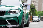 A close up front view of AUTOTECKNIC DRY CARBON PERFORMANTE AERO SPLITTERS - BMW F87 M2 COMPETITION 2018+ fitted on a olive green car