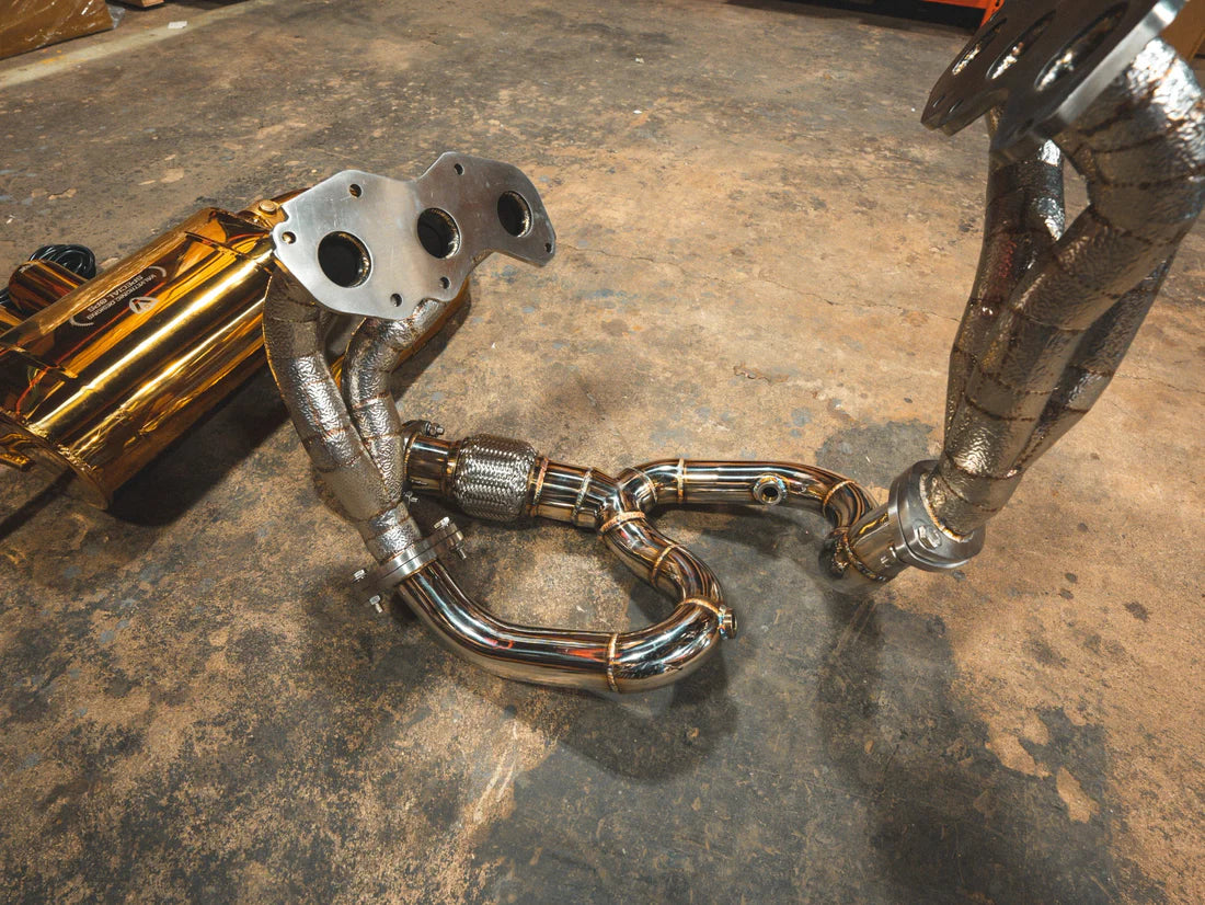 A side view of Valvetronic Designs Anodized Gold Valved Sport Exhaust System's free flow headers on the ground (for Lotus Evora).