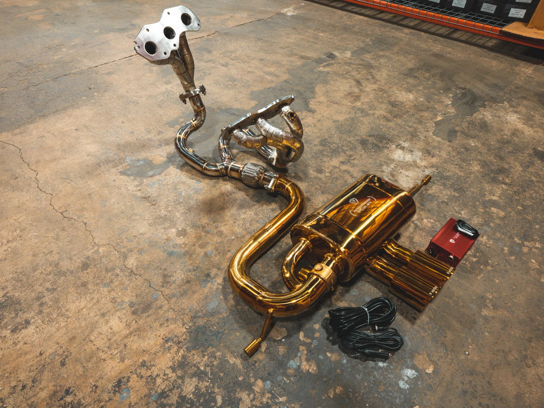 A side overview of Valvetronic Designs Anodized Gold Valved Sport Exhaust System with free flow headers, valve controller and wires on the ground (for Lotus Evora).