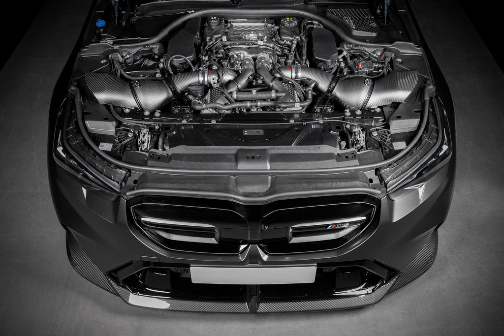 Black carbon fiber turbo inlet tubes installed in BMW M5 engine bay with twin turbo V8 engine visible - Eventuri turbo inlet for BMW G90 G99 M5