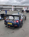 Large black carbon fiber rear wing with multiple support struts mounted on blue race car at track paddock - APR Performance rear wing for Universal 