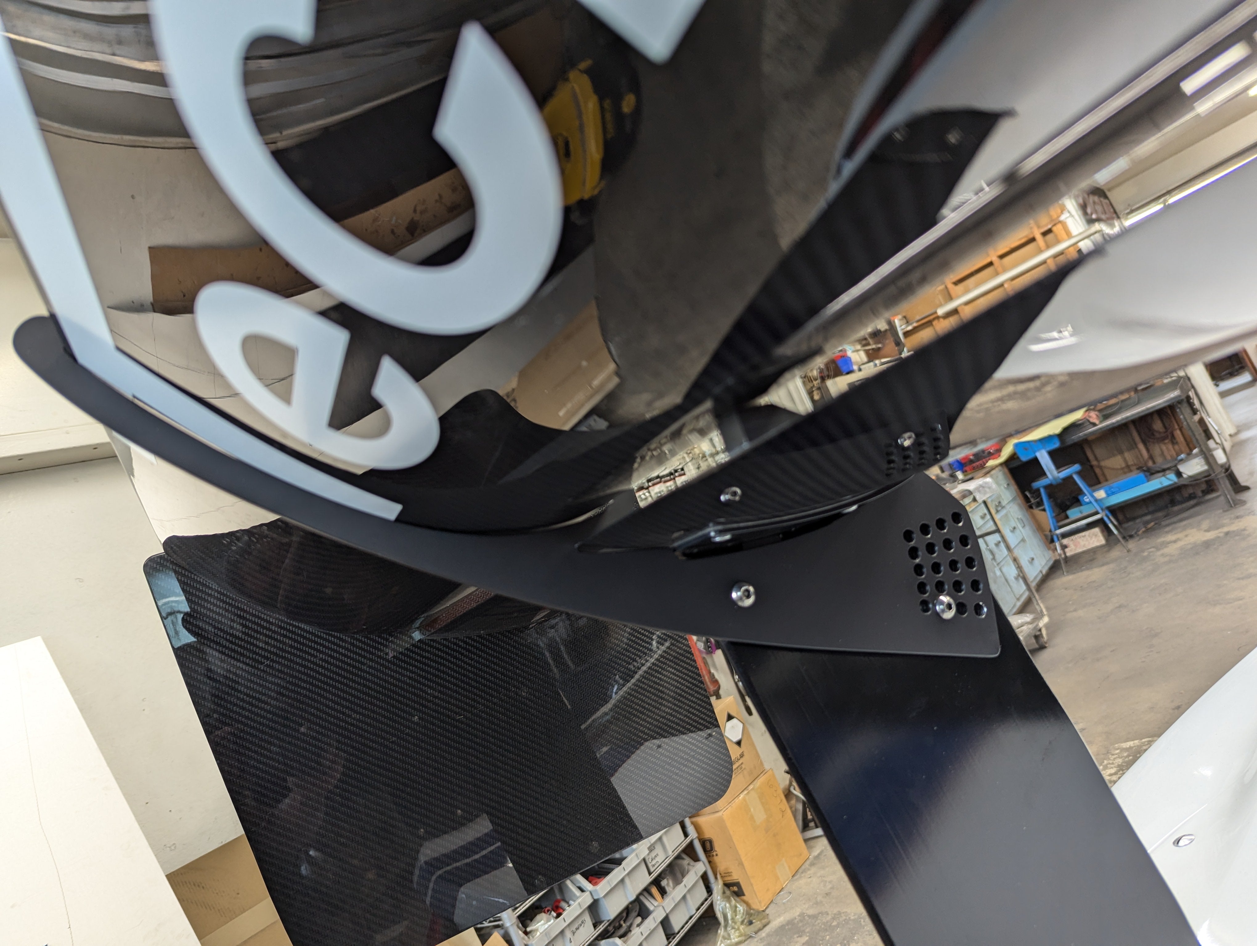 Black carbon fiber rear wing spoiler with adjustable mounting brackets positioned in workshop garage environment - APR Performance rear wing for Universal 