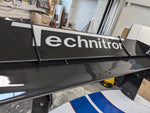 Large carbon fiber rear wing with white lettering displayed in workshop environment with various automotive parts visible - APR Performance rear wing for Universal 