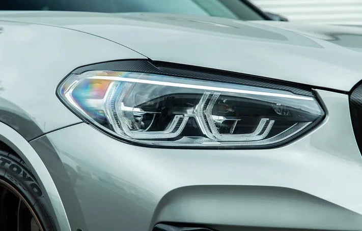 A close up view of AUTOTECKNIC CARBON FIBER HEADLIGHT COVERS - BMW G01 X3 | G02 X4 2017+ fitted on a grey car