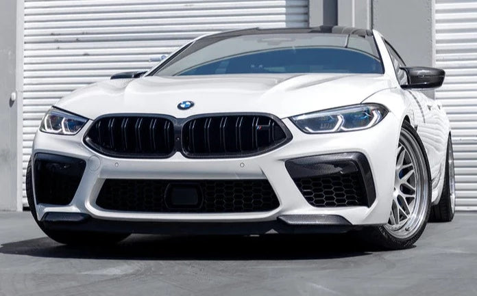 A front view of a white BMW on the road fitted with AUTOTECKNIC PERFORMANCE DRY CARBON SPLITTER SET - BMW F9X M8 2019+