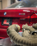 A close up view of RK Autowerks F9X M5 M8 CATLESS DOWNPIPES on a table with a red BMW in the background