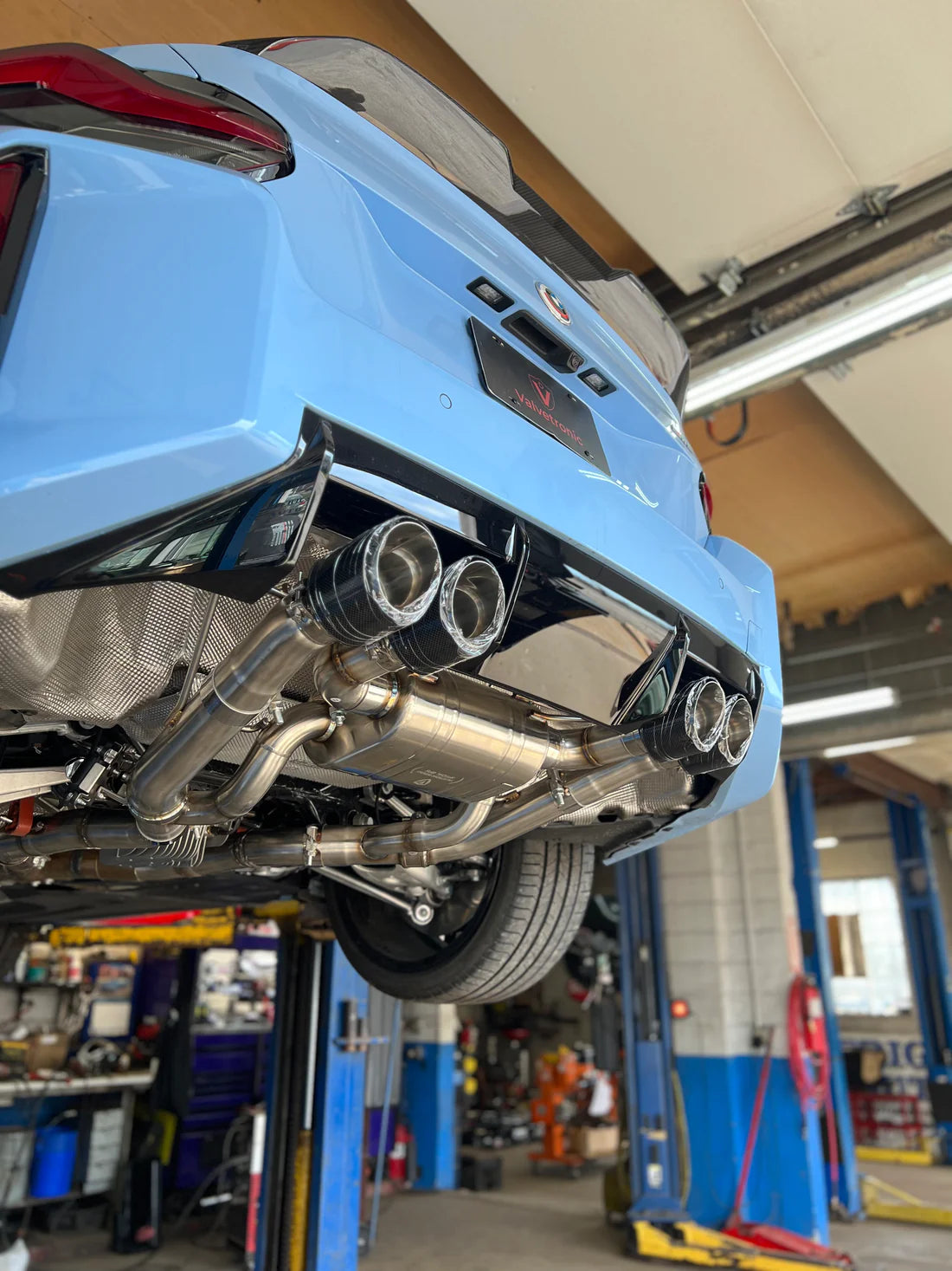 A side view of part of Valvetronic Designs Valved Sport Exhaust System fitted on a lifted blue car from behind (for BMW G87 M2).