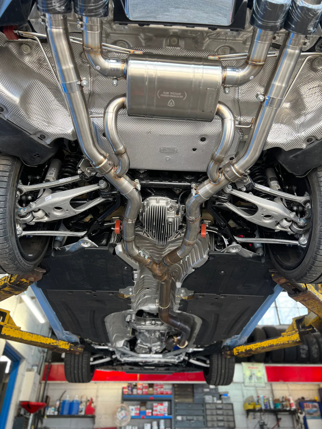 A close view of Valvetronic Designs Valved Sport Exhaust System fitted on a lifted blue car from beneath (for BMW G87 M2). 