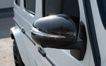 A close up view of AUTOTECKNIC REPLACEMENT DRY CARBON MIRROR COVERS - MERCEDES-BENZ W463A G-WAGON 2019+ fitted on a white car outside on the road